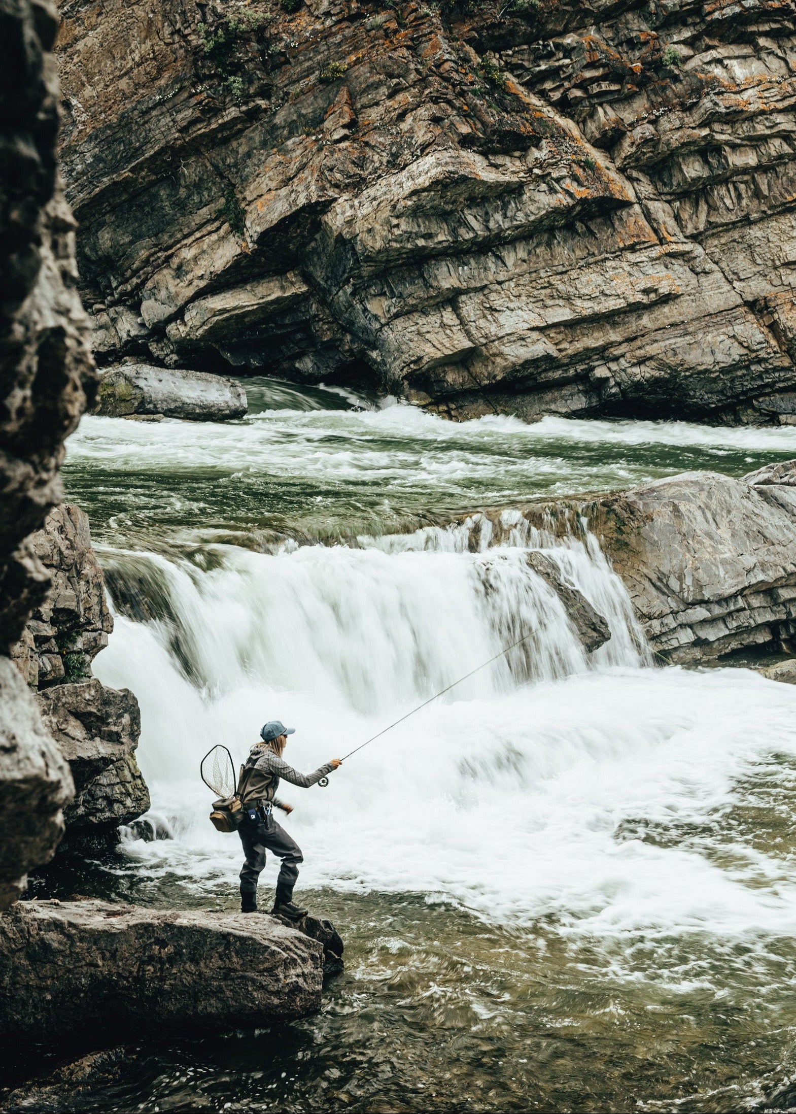 A person fly fishing on a rocky riverbank with rushing water and steep cliffs in the background.