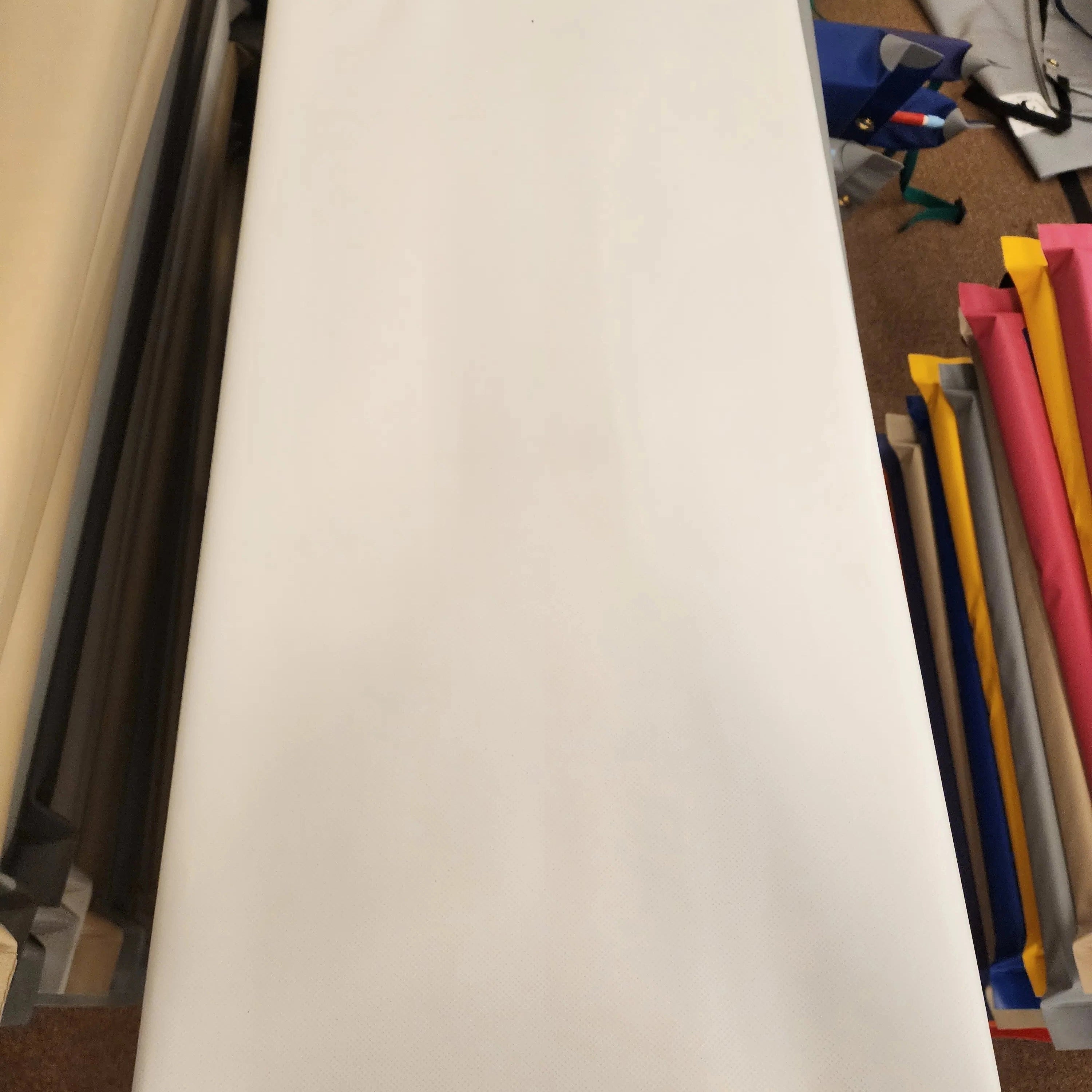 A large white canvas banner or flag with brass grommets on the left and right edges, featuring a small green square logo patch on the bottom left, photographed in a workshop or storage area with other colorful rolled materials in the background.