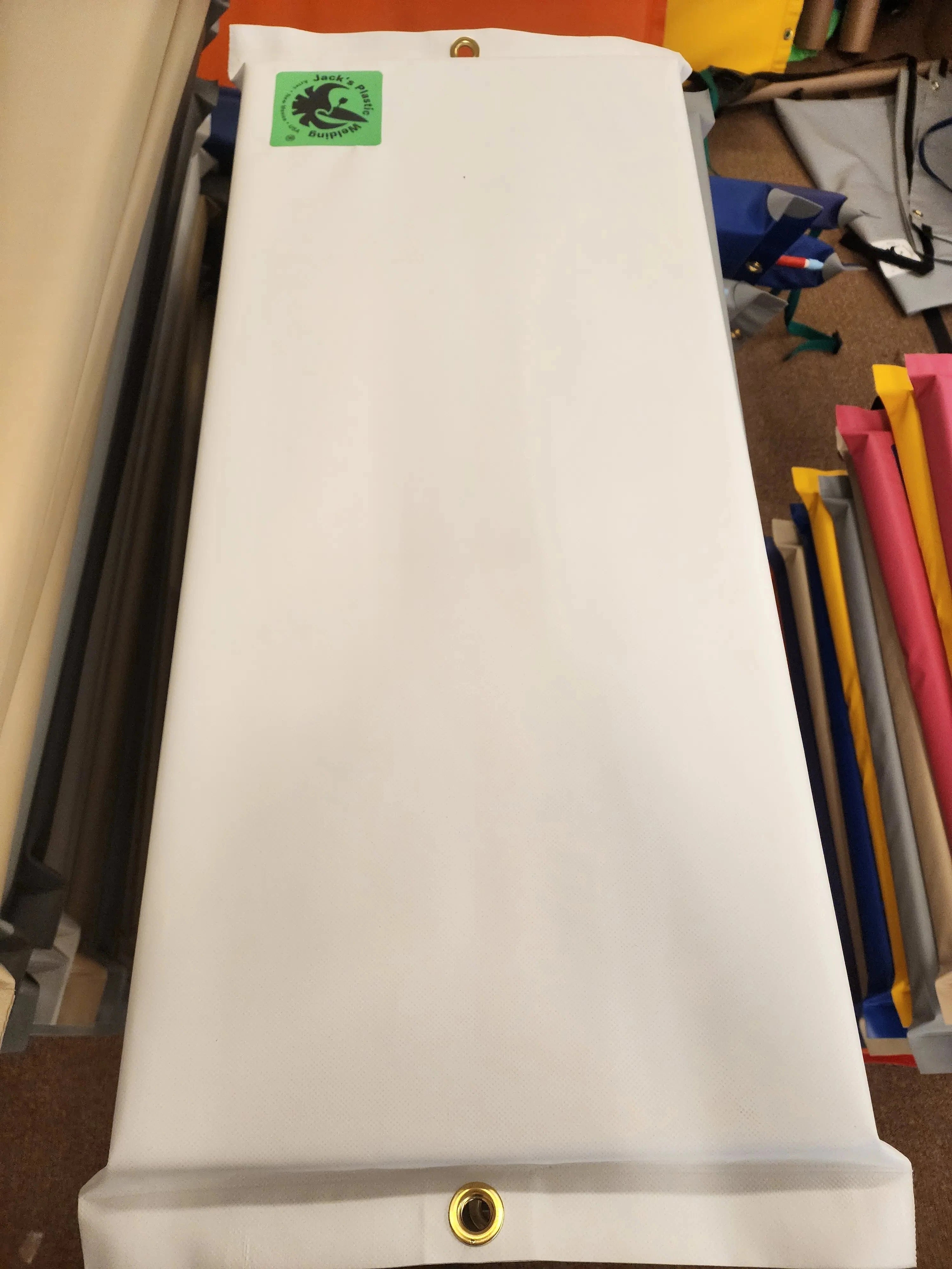 A large white canvas banner or flag with brass grommets on the left and right edges, featuring a small green square logo patch on the bottom left, photographed in a workshop or storage area with other colorful rolled materials in the background.
