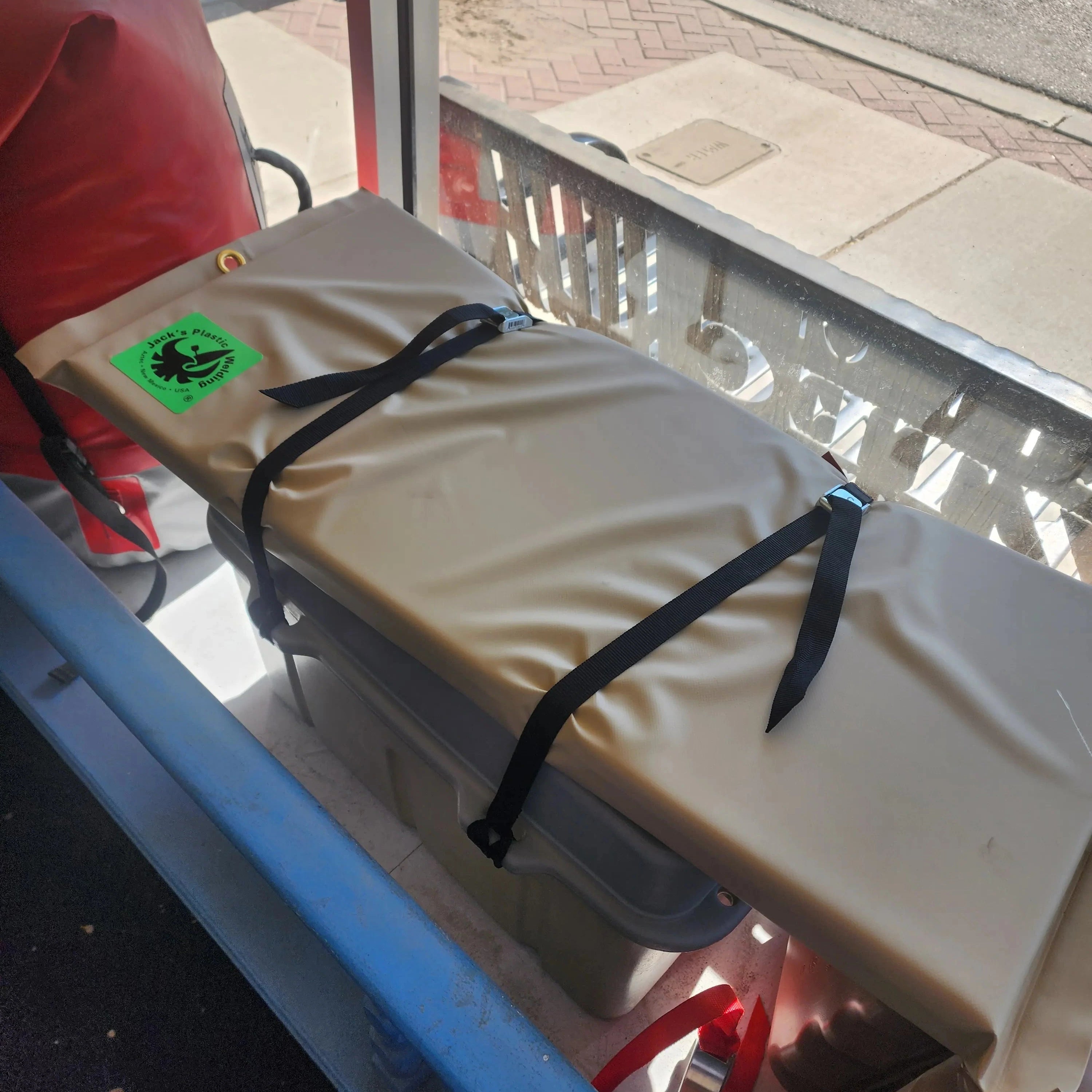 A beige padded rectangular item secured with black straps, photographed in a real environment near a window and blue frame, with a small green sticker visible on the lower left.