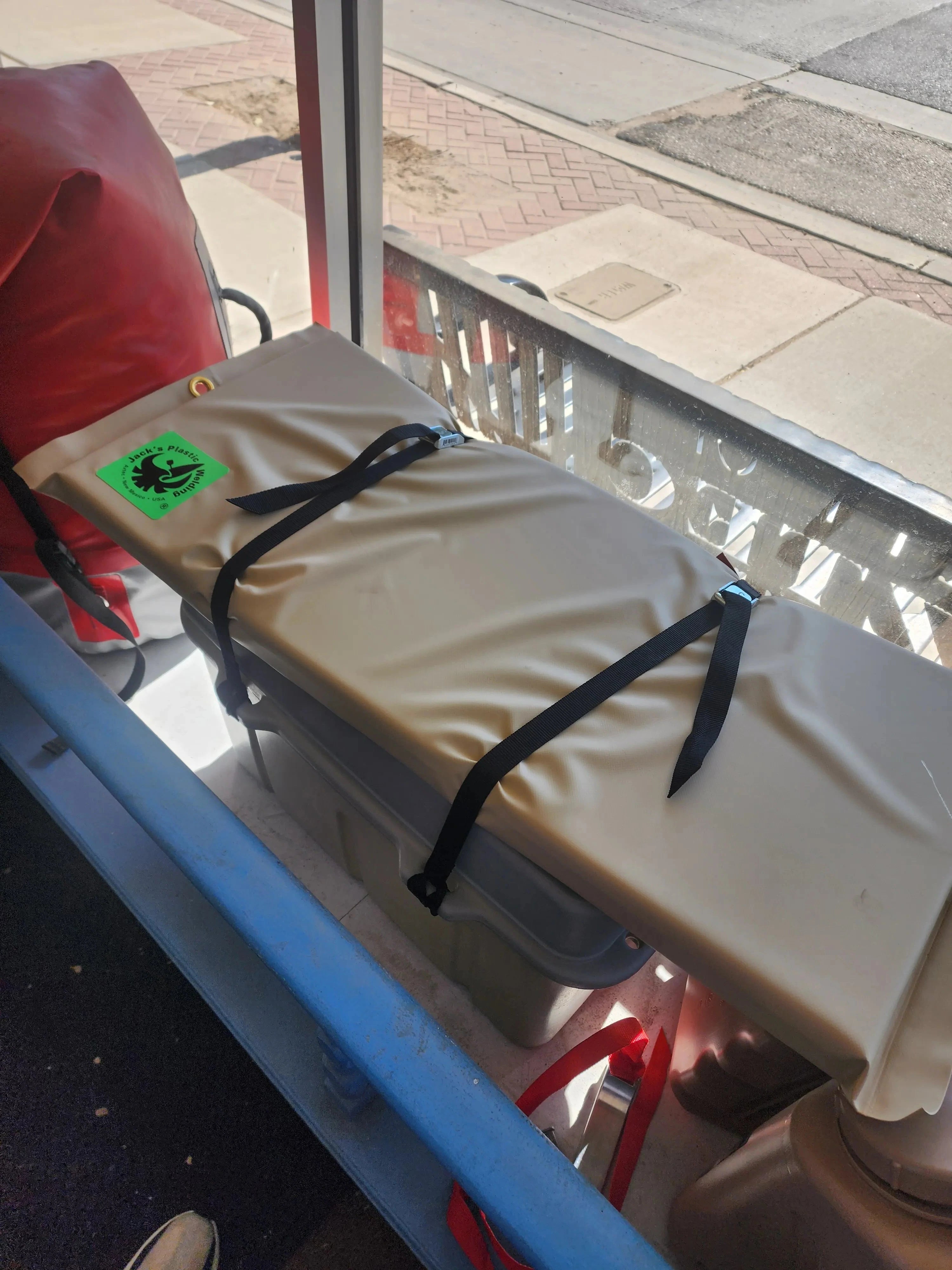 A beige padded rectangular item secured with black straps, photographed in a real environment near a window and blue frame, with a small green sticker visible on the lower left.