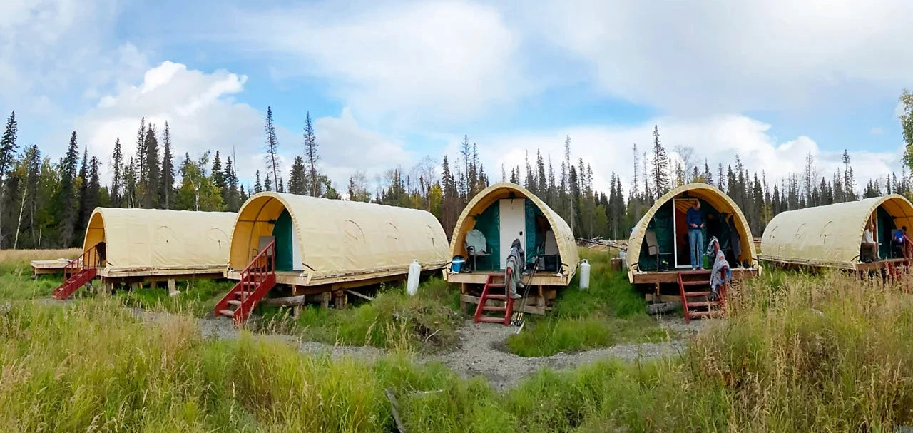Four dome-shaped glamping pods with cream fabric walls and red wooden stairs, set in a grassy field with a forest and blue sky in the background.