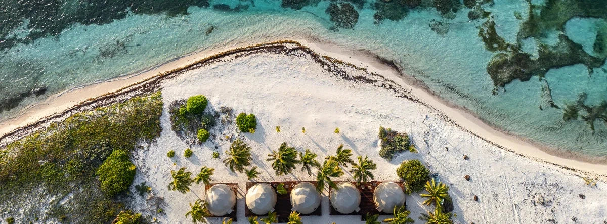 Aerial view of a tropical beach with turquoise water, white sand, palm trees, and a row of dome-shaped beach huts on the shore.