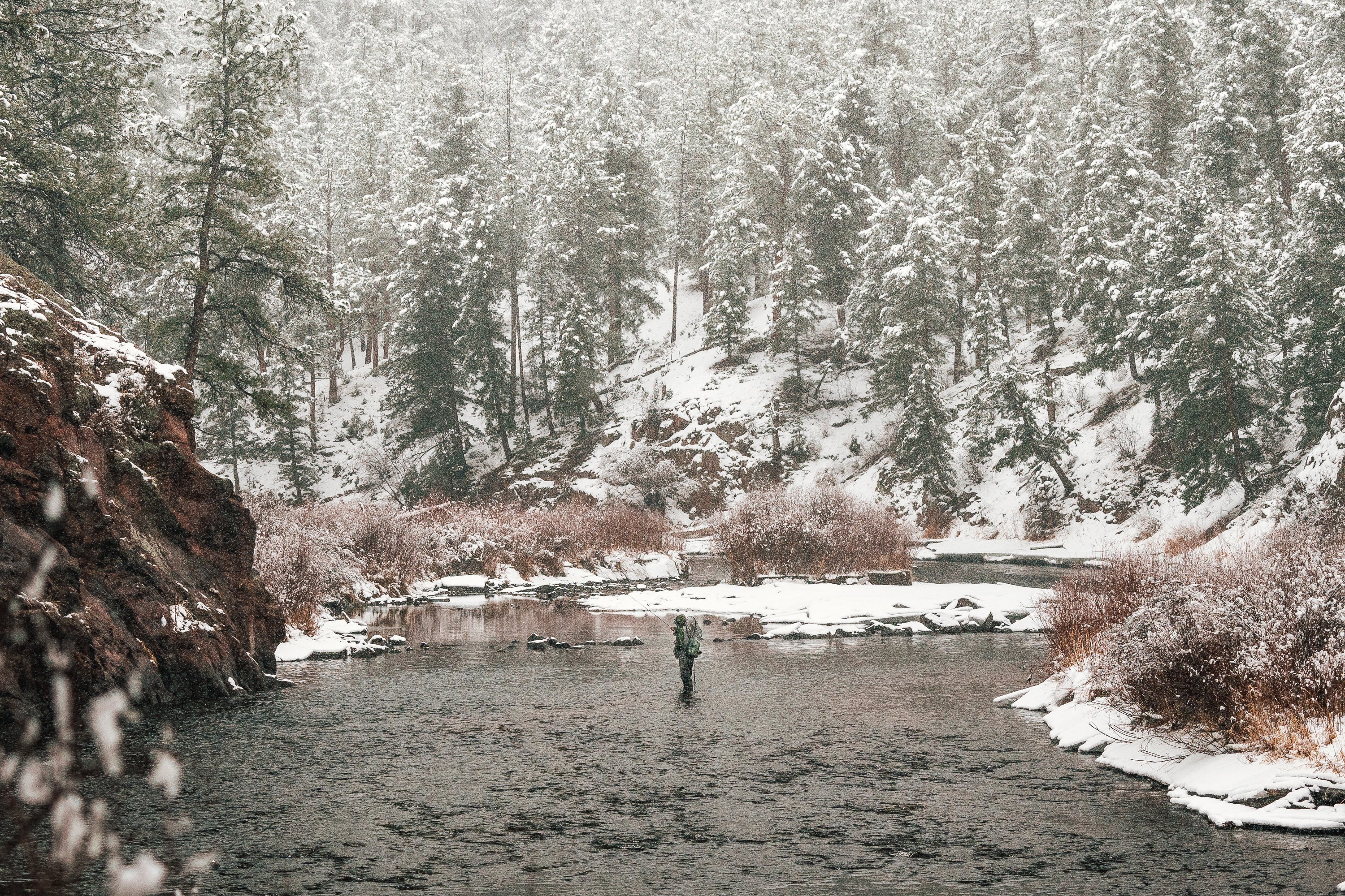 Person fishing in a river surrounded by snow-covered trees and rocks.