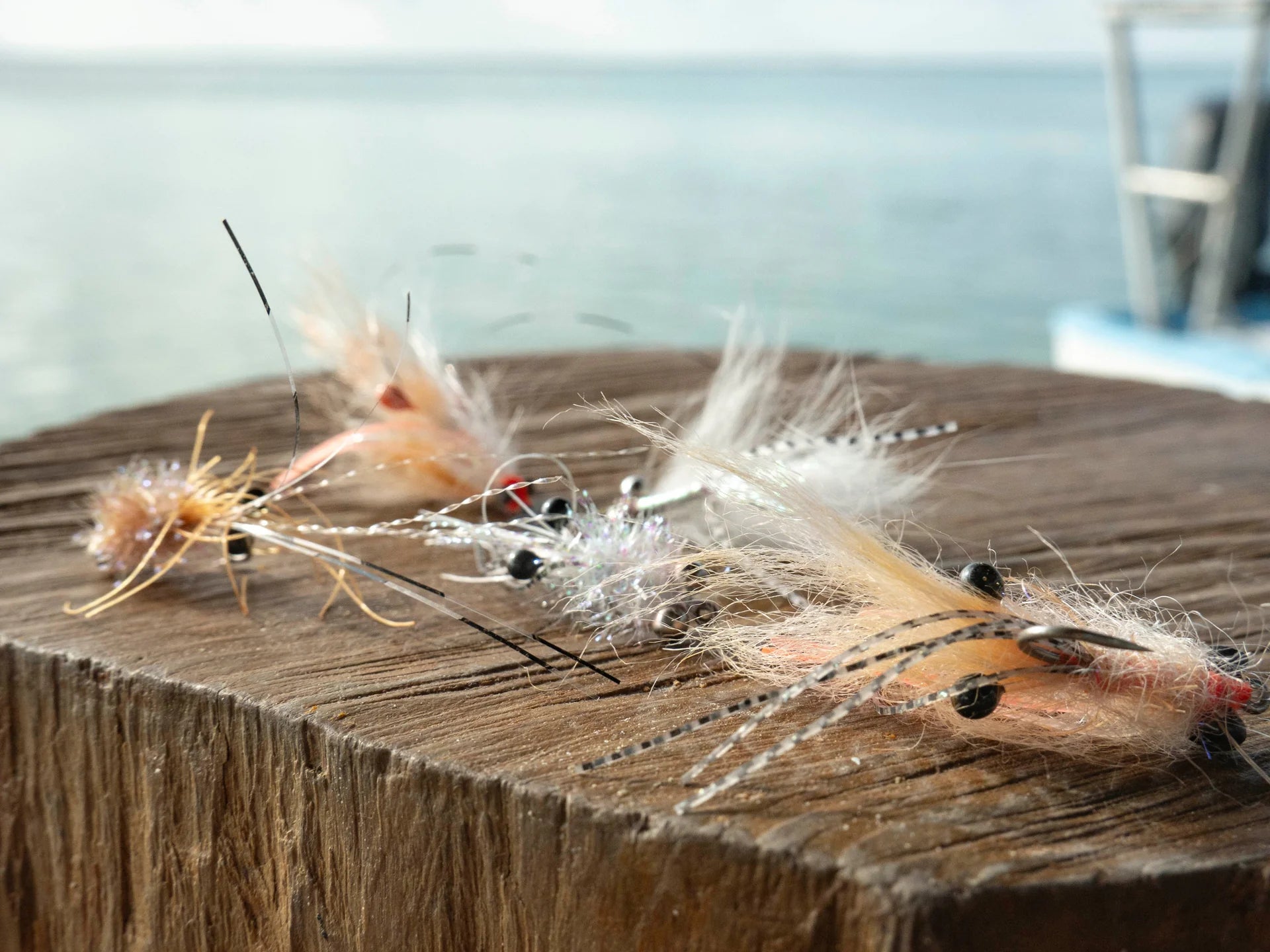 Three fly fishing lures with feathers and thread resting on the edge of a wooden dock, with a blurred body of water and a boat in the background.