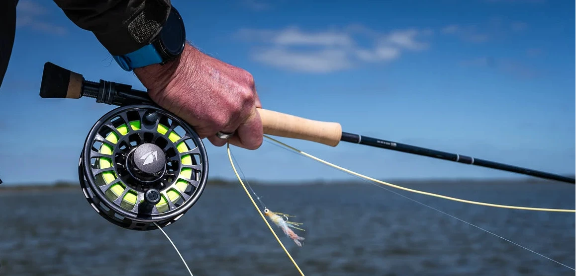 A close-up of a person's hand holding a fly fishing rod with a black and green fly reel, casting line over a body of water under a blue sky.