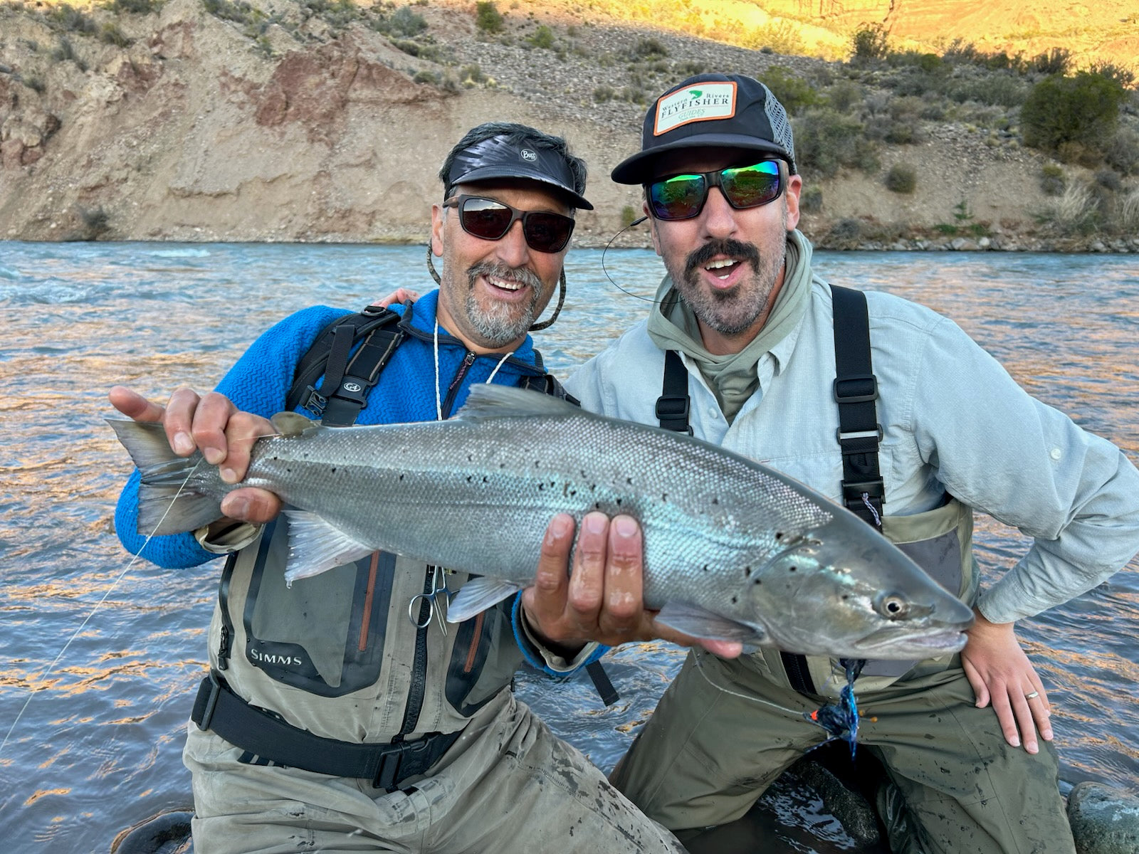 Two men in fly-fishing gear and sunglasses pose with a large salmon on a river, with a rocky bank in the background.