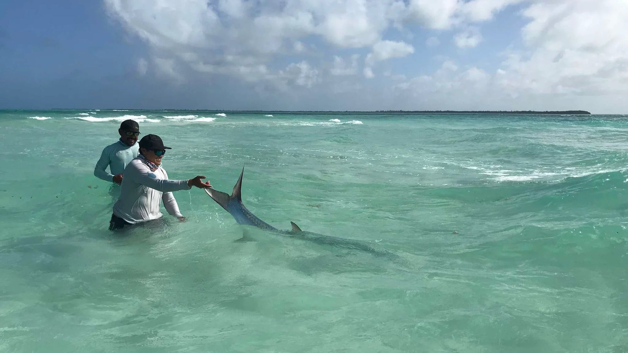 Two men standing in shallow turquoise ocean water hold a large shark by its tail fins under a cloudy blue sky.