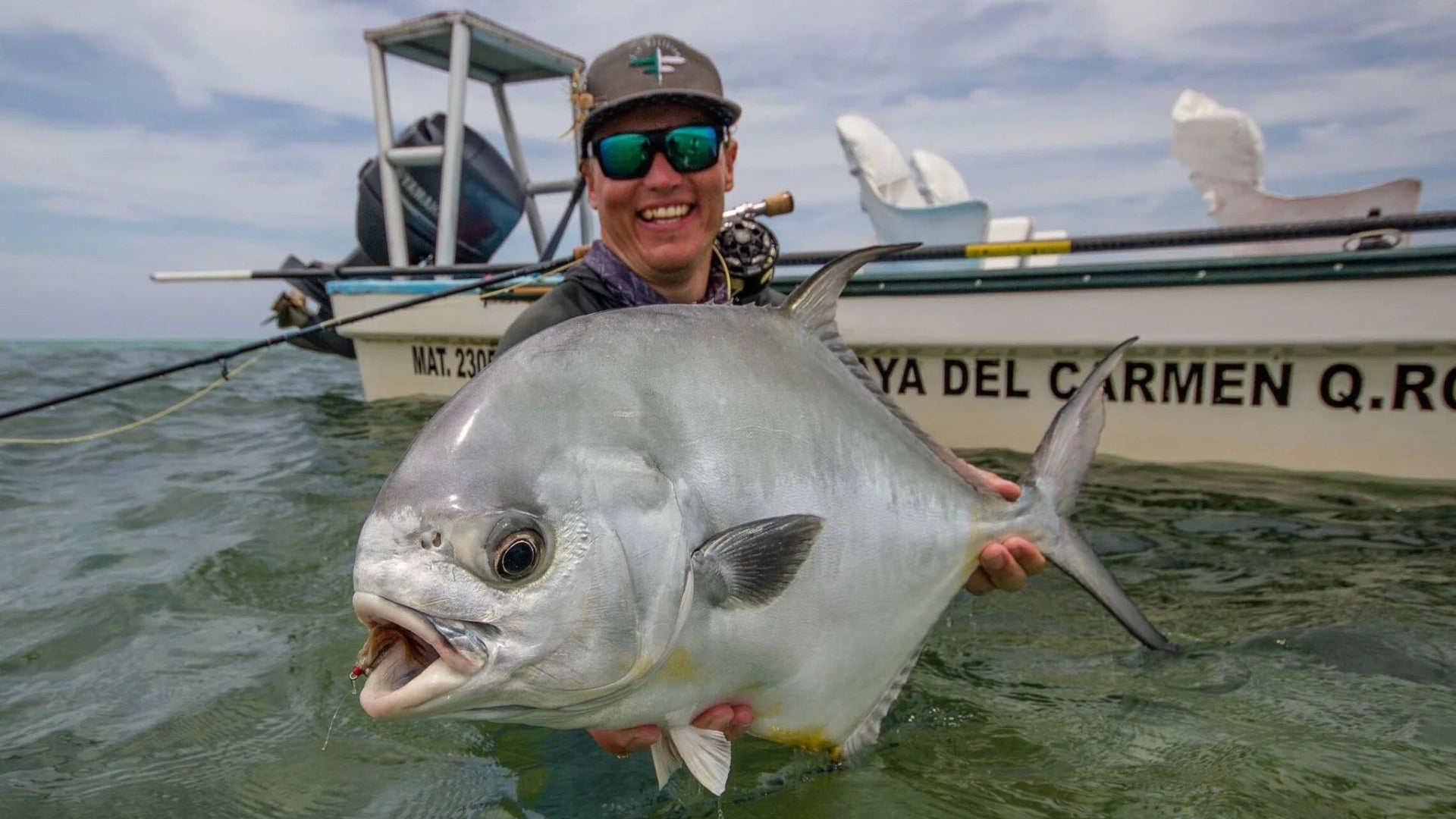 A smiling man in a cap and mirrored sunglasses holds a large silver fish in shallow water, with a boat visible in the background under a cloudy sky.