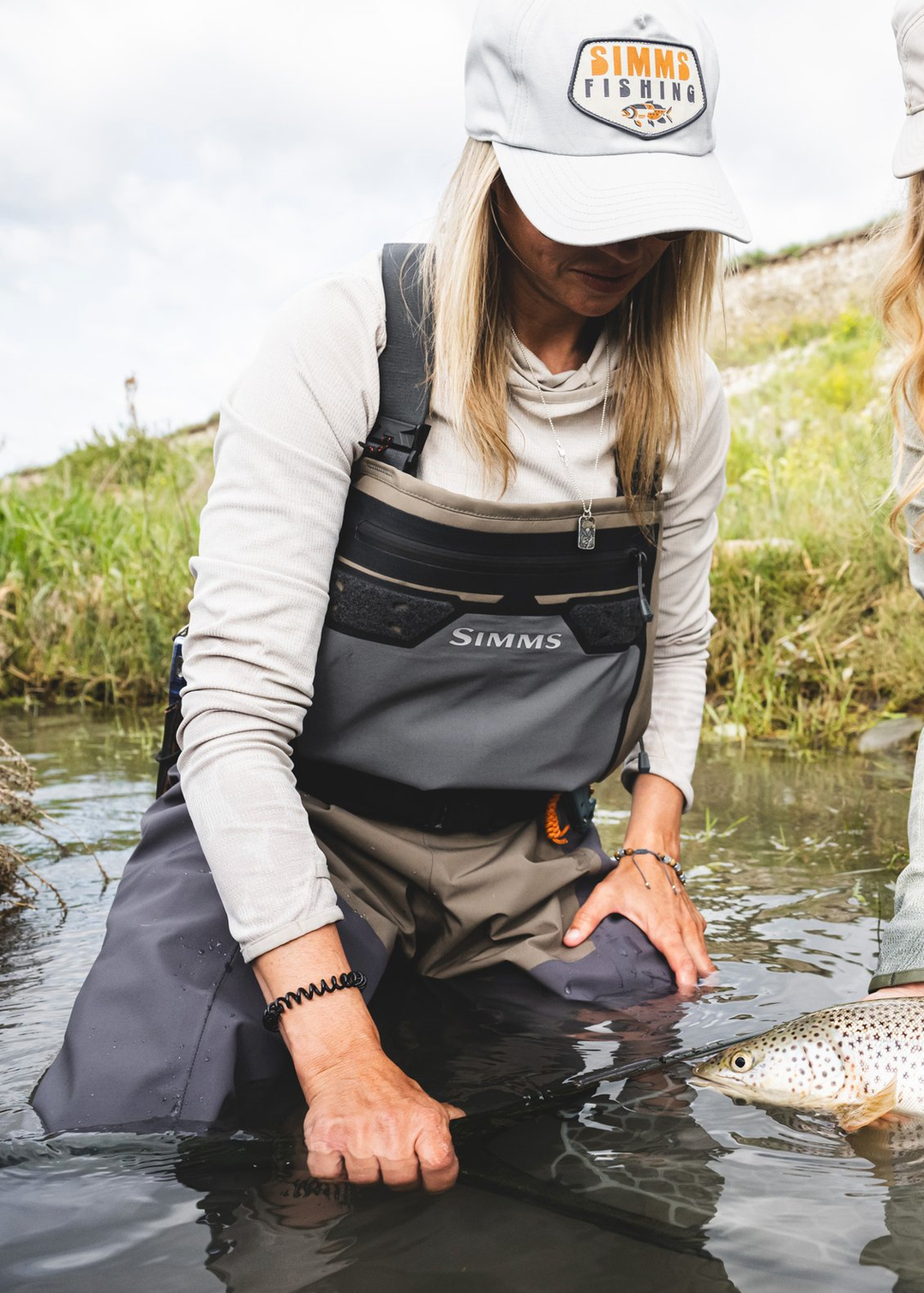 Two women in Simmo waders and fishing gear kneel in a river, holding a large brown trout. They wear matching gray and black Simmo waders and white Simmo caps, with a fly rod visible in the water.