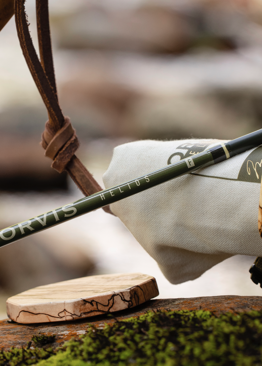 A close-up of an Orvis fly fishing rod resting on a mossy log, with its wooden case partially visible and a leather loop nearby, set against a blurred outdoor background.