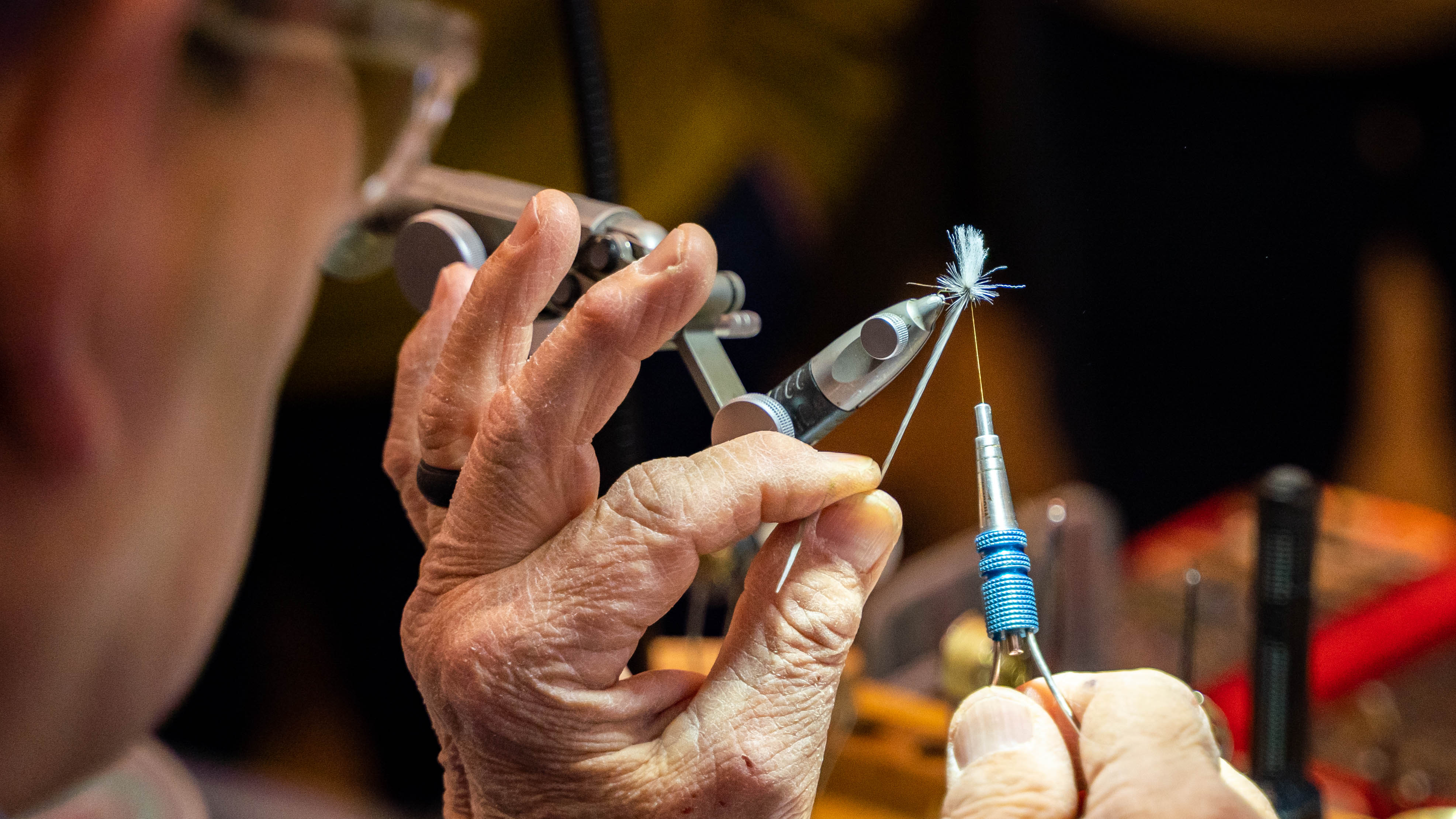 A close-up lifestyle shot of a fly tying artisan's hands using specialized tools, including forceps and a thread-winder, to craft a small blue and white fishing fly. The background is a softly blurred workshop environment.
